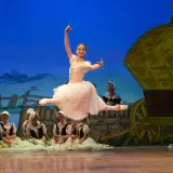 Baletnica w skoku, główna postać pięknej choreografii. W tle wiejska scenografia z wozem i innymi tancerzami.