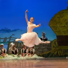 Baletnica w skoku, główna postać pięknej choreografii. W tle wiejska scenografia z wozem i innymi tancerzami.