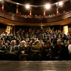 Widownia w teatrze, ludzie siedzą na miejscach, widać balkon z widzami.