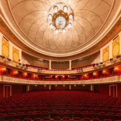 Wnętrze teatru z czerwonymi fotelami i bogato zdobionym sufitem. Czekamy na wspaniały występ.