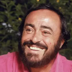 Uśmiechnięty Luciano Pavarotti w różowej koszuli, z charakterystyczną brodą i bujnymi włosami.