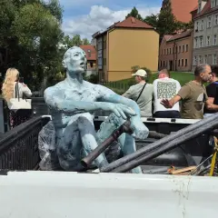 Rzeźbiarze transportują niebieską rzeźbę siedzącego mężczyzny w bagażniku pickupa. W tle widać budynki i ludzi.
