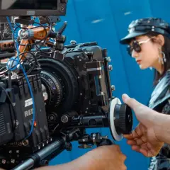 Ekipa filmowa pracuje nad sesją zdjęciową. W centrum uwagi profesjonalna kamera, a w tle modelka w stylowej garderobianej stylizacji.