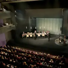 Scena teatralna z ludźmi przy stole i instrumentami muzycznymi. Widownia siedzi w ciemności, gotowa na spektakl. Dostawka w teatrze.
