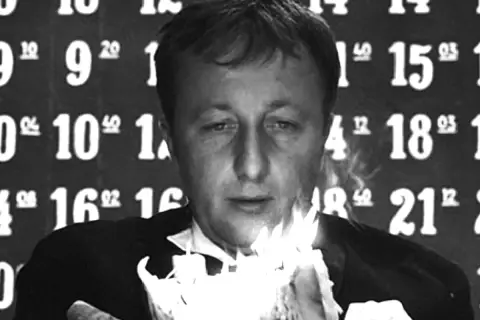 Mężczyzna w garniturze trzyma płonące świece. Tło to liczby, jakby czas się zatrzymał, symbolizując cenzurę i przemijanie.