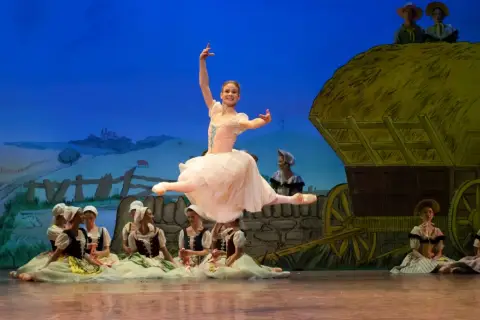 Baletnica w skoku, główna postać pięknej choreografii. W tle wiejska scenografia z wozem i innymi tancerzami.
