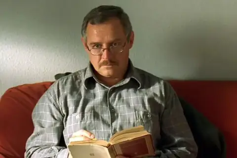 Mężczyzna w okularach i kratowanej koszuli czyta książkę. To Marek Kondrat, jeden z aktorów z obsady "Dnia świra".