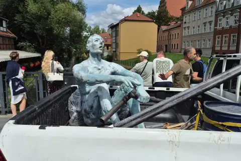 Rzeźbiarze transportują niebieską rzeźbę siedzącego mężczyzny w bagażniku pickupa. W tle widać budynki i ludzi.