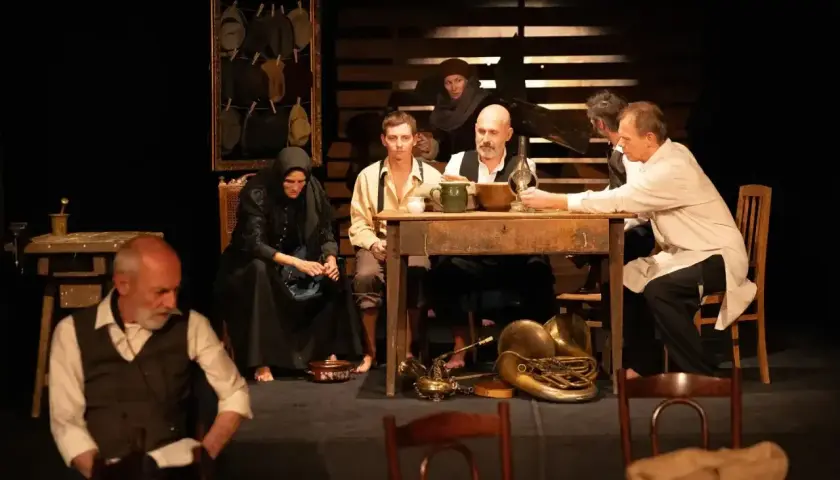 Scena teatralna, grupa ludzi przy stole, atmosfera napięcia i oczekiwania. Ten dramat porusza trudne tematy.