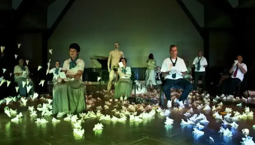 Scena teatralna pełna papierowych ptaków, ludzie w strojach z epoki i współczesnych ubraniach, posąg. To wszystko tworzy atmosferę teatru absurdu.
