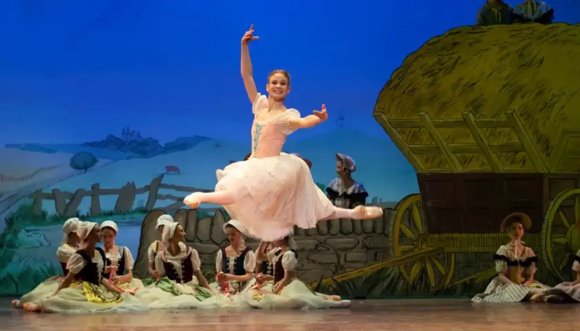 Baletnica w skoku, główna postać pięknej choreografii. W tle wiejska scenografia z wozem i innymi tancerzami.