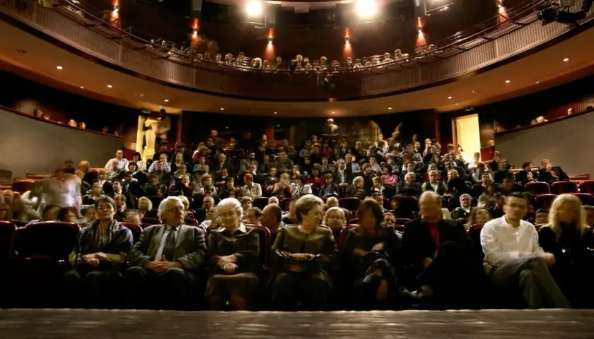 Widownia w teatrze, ludzie siedzą na miejscach, widać balkon z widzami.