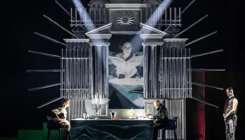 Scena teatralna z projekcją twarzy kobiety, dwójką siedzących przy stole postaci i stojącym mężczyzną. Atmosfera pełna jest teatralnych misteriów.