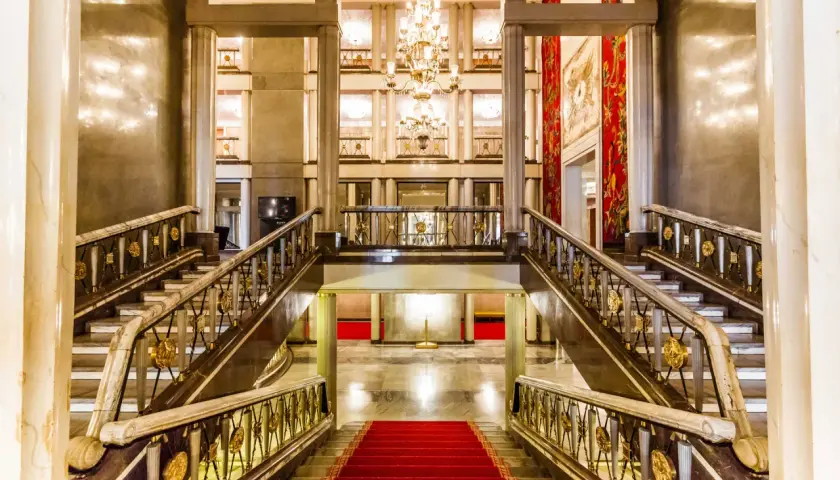 Elegancki foyer z marmurowymi schodami, złotymi balustradami i kryształowym żyrandolem. Czerwony dywan prowadzi do przestronnego holu.