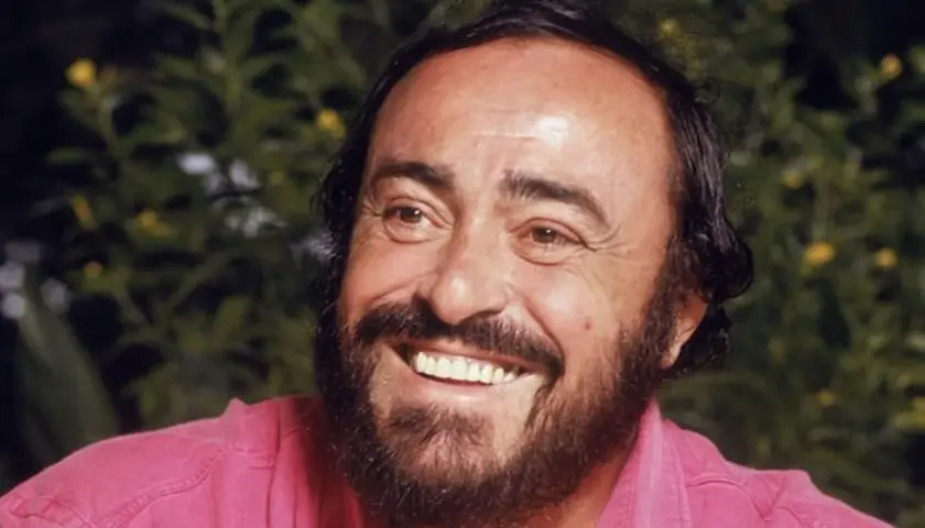Uśmiechnięty Luciano Pavarotti w różowej koszuli, z charakterystyczną brodą i bujnymi włosami.