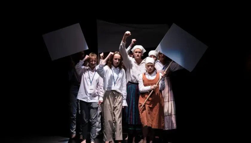 Młoda grupa teatralna z transparentami wznosi pięści. Występ na czarnym tle.