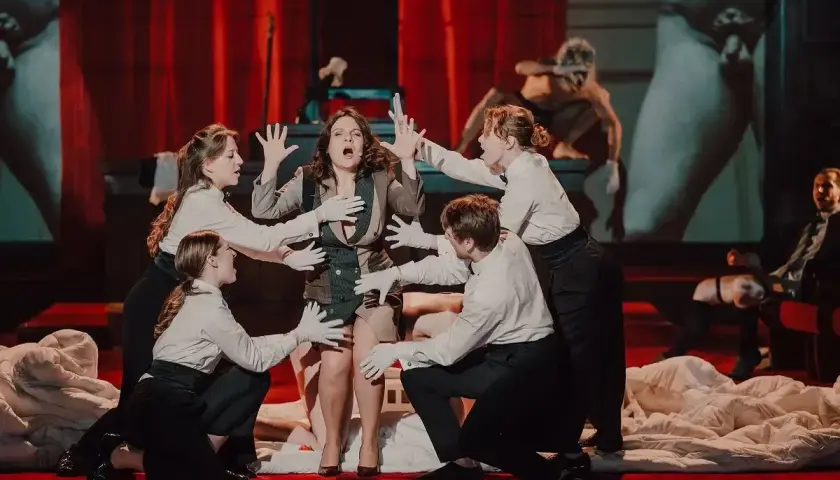 Scena z przedstawienia teatralnego, gdzie aktorzy w białych rękawiczkach otaczają kobietę. To jeden z wielu emocjonujących spektakli, jakie oferują teatry w Polsce.