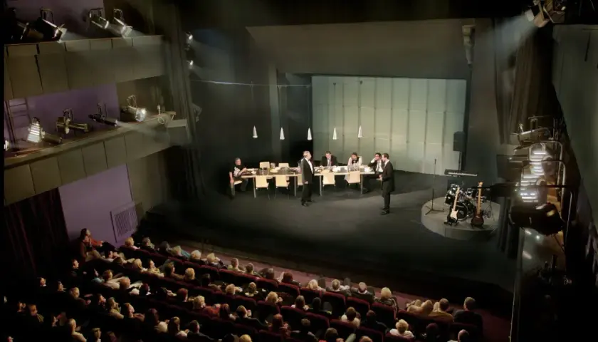 Scena teatralna z ludźmi przy stole i instrumentami muzycznymi. Widownia siedzi w ciemności, gotowa na spektakl. Dostawka w teatrze.