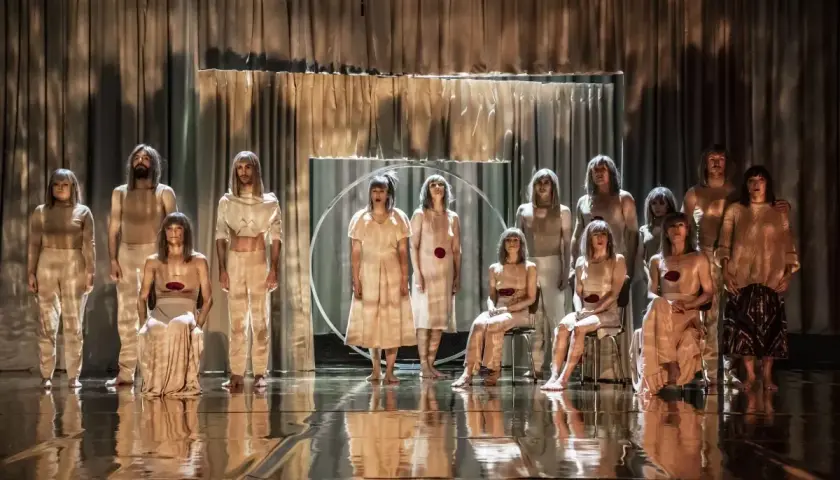 Grupa ludzi w beżowych strojach, jakby w scenicznej pantomimie, stoi na błyszczącej scenie.