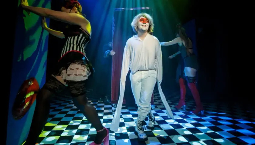 Scena teatralna, gdzie młody mężczyzna w białym stroju i makijażu twarzy stoi pośród postaci w kostiumach, tworząc atmosferę tragifarsy.