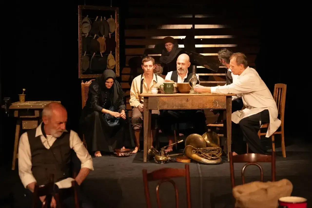 Scena teatralna, grupa ludzi przy stole, atmosfera napięcia i oczekiwania. Ten dramat porusza trudne tematy.