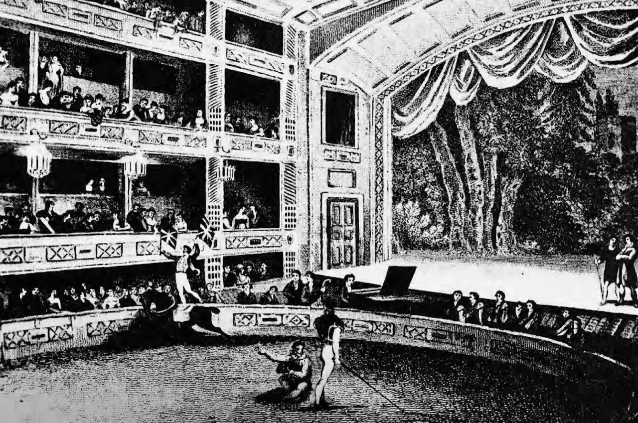 Widownia wypełnia wielopoziomowe loże, a na arenie trwa spektakl. To scena z życia teatru średniowiecznego, pełna widzów i aktorów.