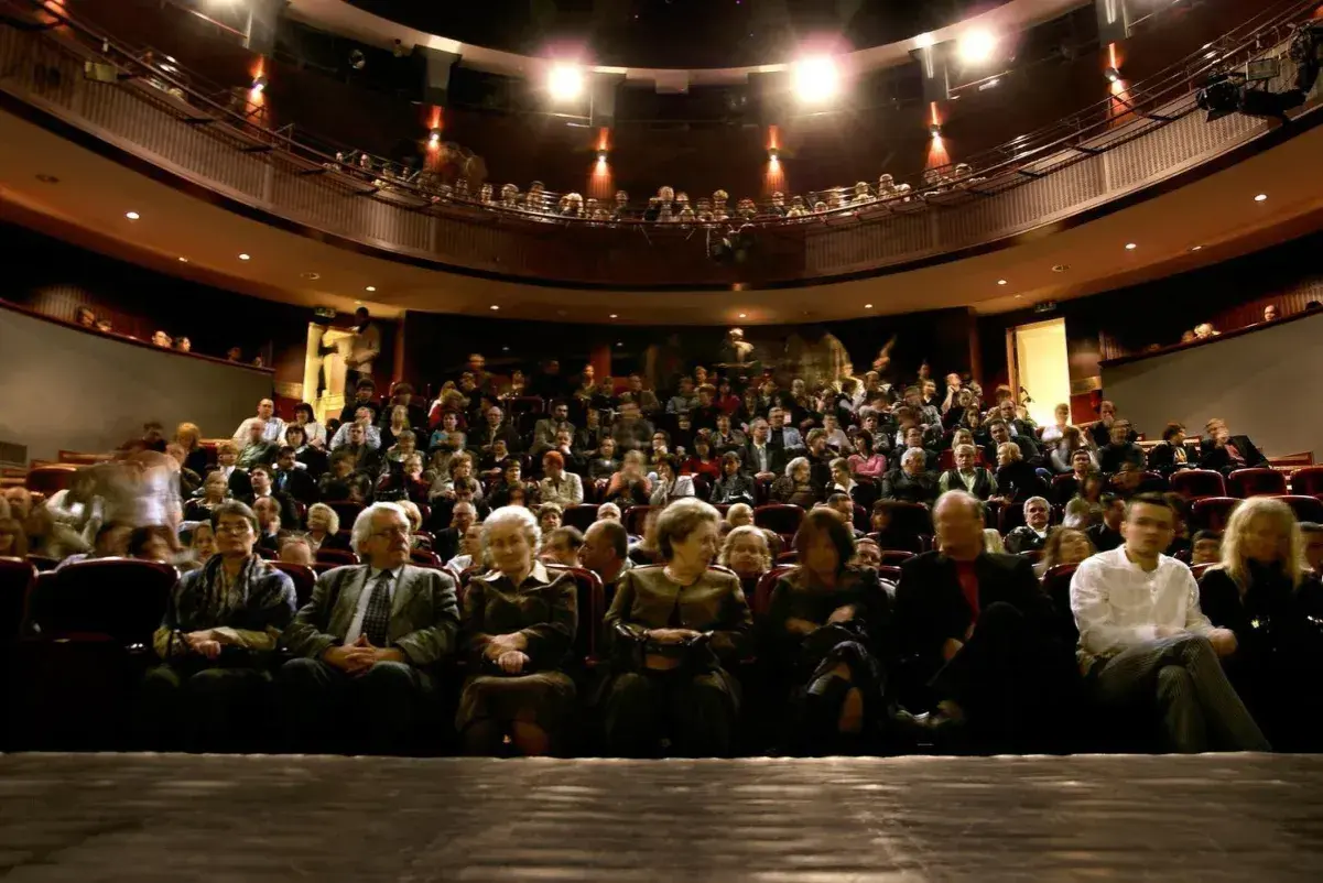 Widownia w teatrze, ludzie siedzą na miejscach, widać balkon z widzami.