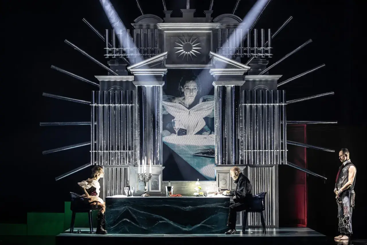 Scena teatralna z projekcją twarzy kobiety, dwójką siedzących przy stole postaci i stojącym mężczyzną. Atmosfera pełna jest teatralnych misteriów.