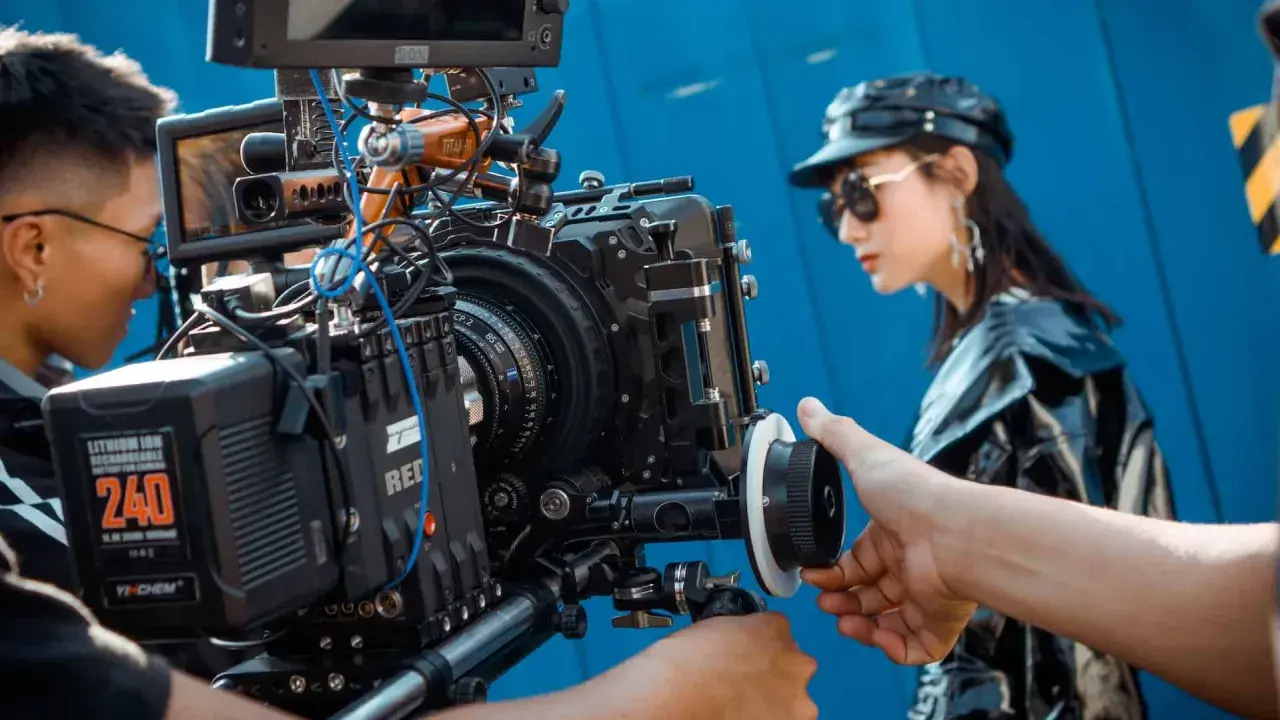 Ekipa filmowa pracuje nad sesją zdjęciową. W centrum uwagi profesjonalna kamera, a w tle modelka w stylowej garderobianej stylizacji.