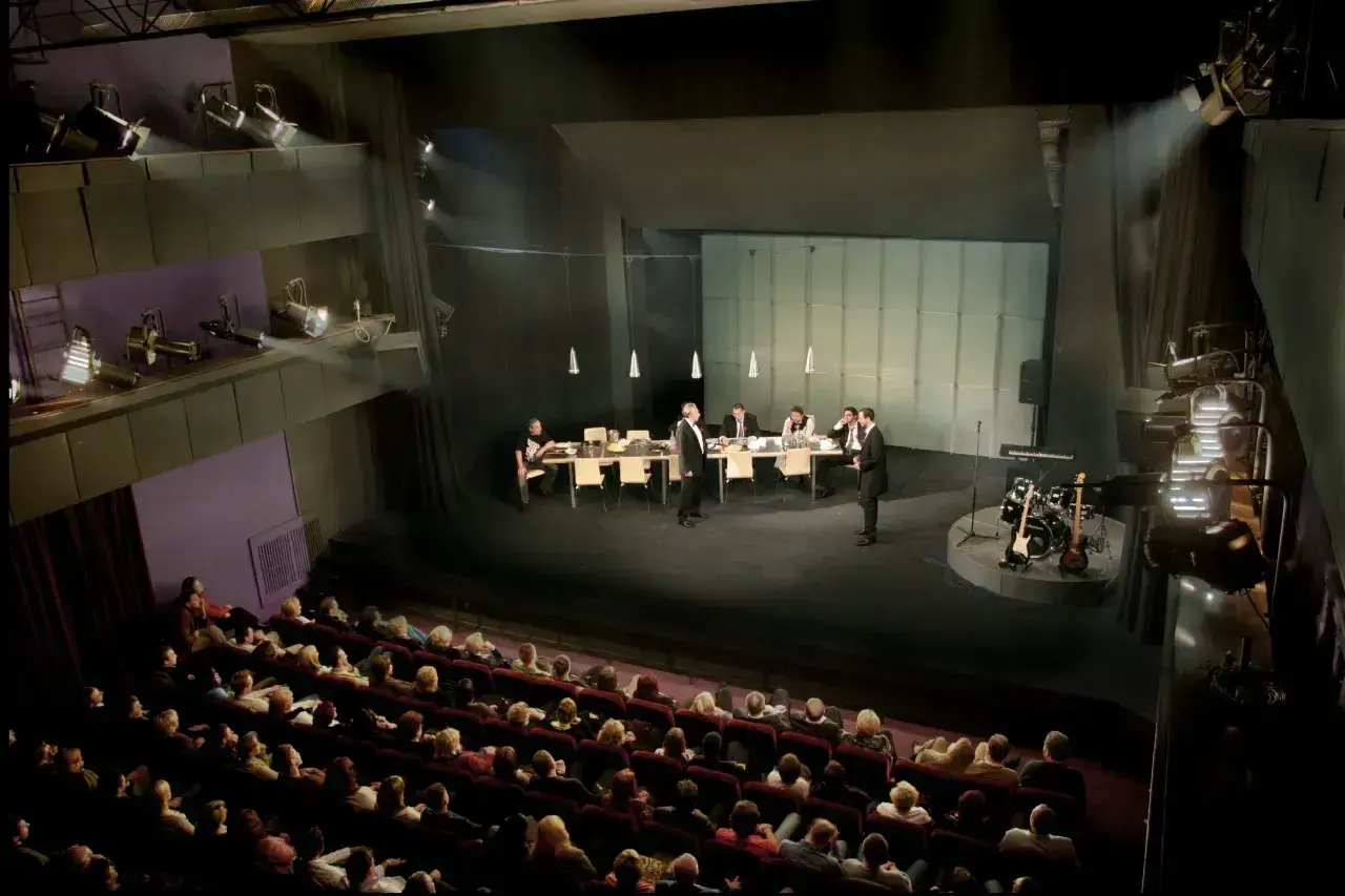 Scena teatralna z ludźmi przy stole i instrumentami muzycznymi. Widownia siedzi w ciemności, gotowa na spektakl. Dostawka w teatrze.