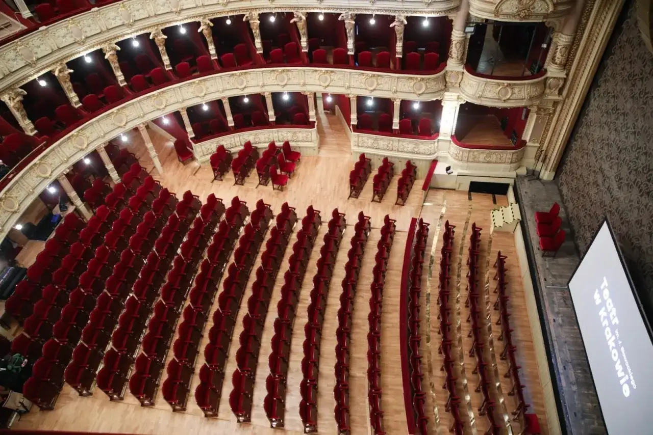 Widok z góry na pustą salę teatralną z rzędami czerwonych foteli. Widać loże i balkon. Na scenie ekran z napisem 