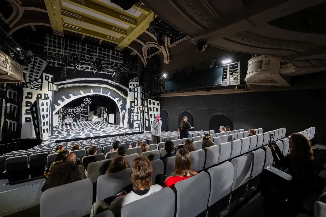 Widownia w teatrze, przed sceną z czarno-białą scenografią. Dwóch aktorów rozmawia z publicznością.