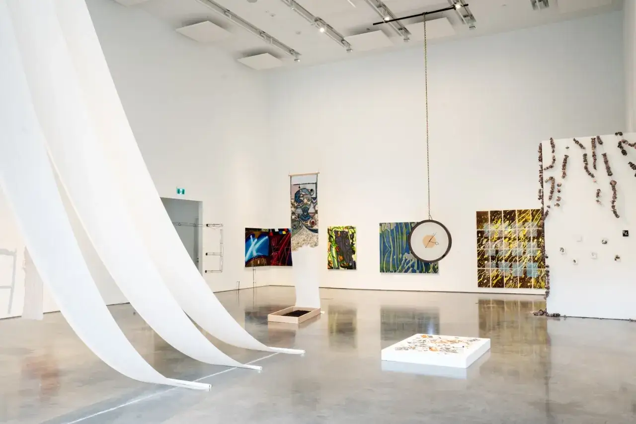 Przestronna galeria sztuki z nowoczesną instalacją artystyczną. Białe tkaniny spływają z sufitu, a na ścianach wiszą kolorowe obrazy i obiekty.