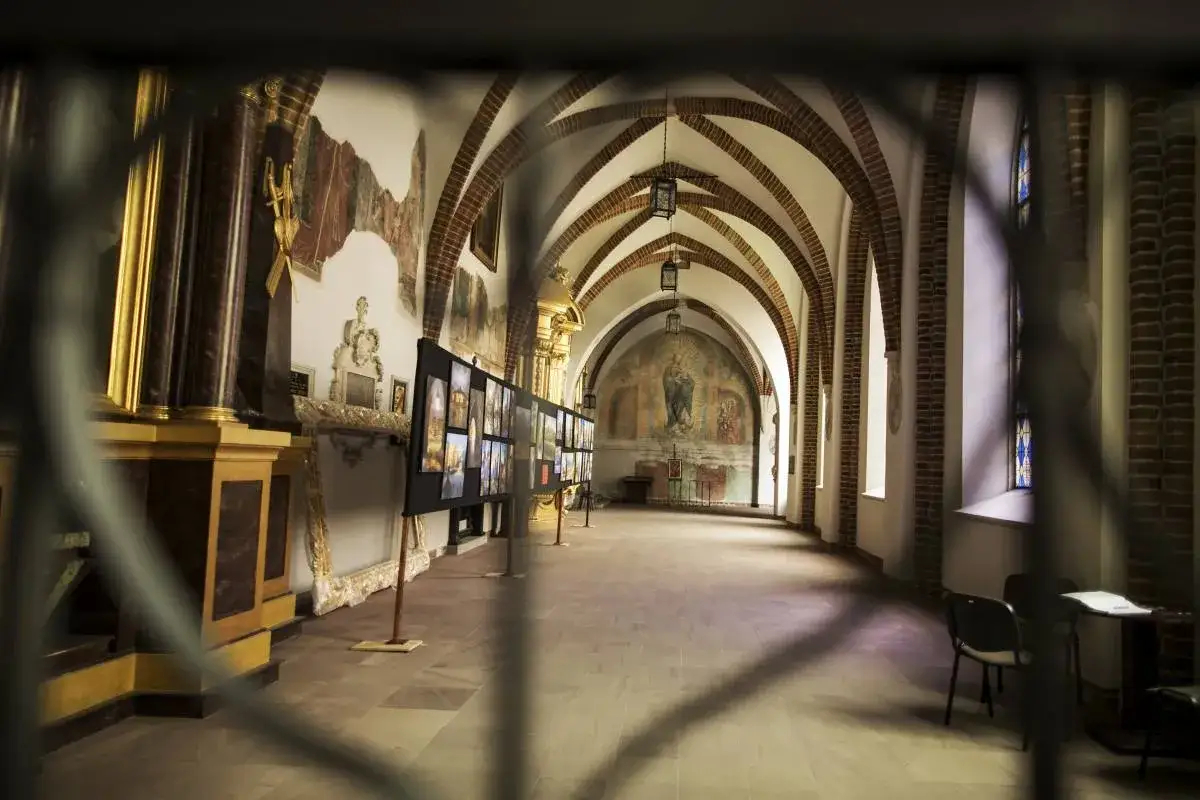 Wnętrze kościoła z arkadami i freskami, przypominające scenografię dla średniowiecznego teatru. Wystawa fotografii na sztalugach.