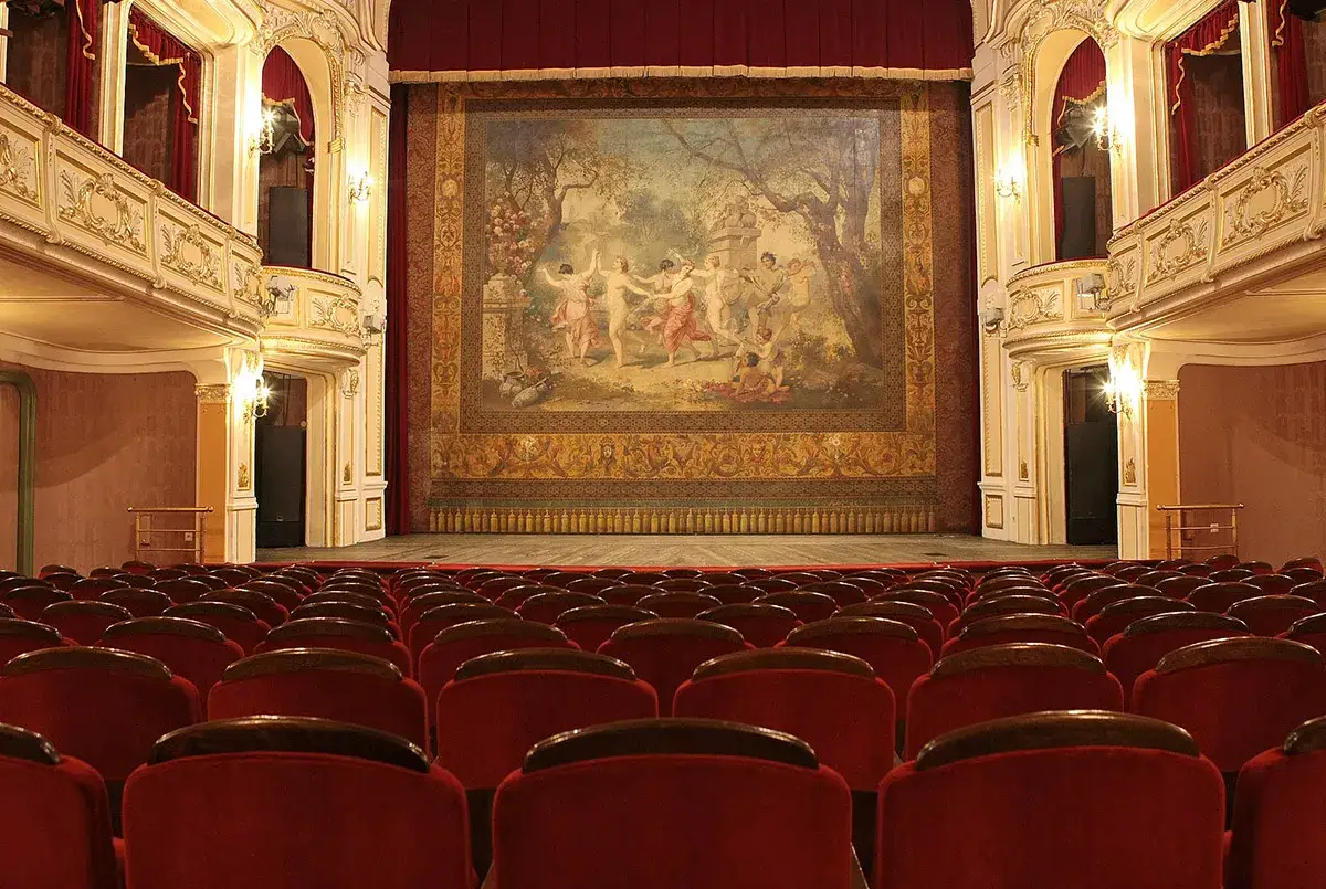 Widok na scenę i rzędy czerwonych foteli. Złocony balkon w teatrze ozdobiony jest malowidłem.
