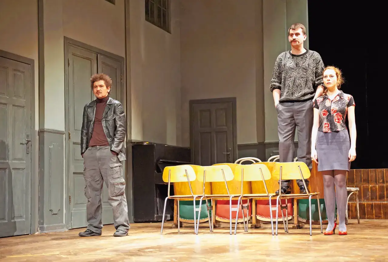 Tragifarsa kołtuńska na scenie. Trzech aktorów w teatralnych strojach, za nimi rząd żółtych krzeseł i drzwi.